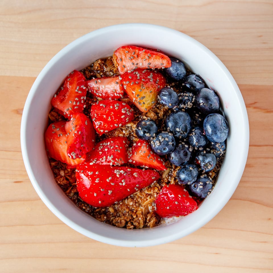 Overhead view of a white bowl filled with granola, strawberries, blueberries, and chia seeds.