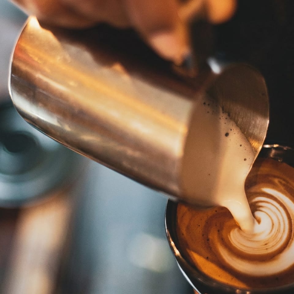 Barista pouring steamed milk from a metal pitcher into a coffee cup, creating latte art.
