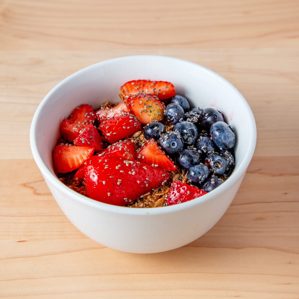 A white bowl filled with granola, fresh strawberries, blueberries, and chia seeds on a wooden table.