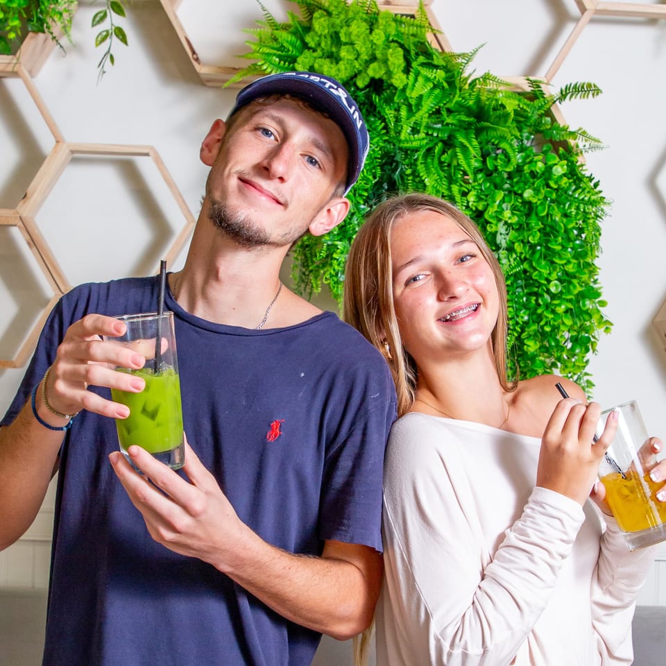 Two smiling young people holding drinks, a green one and a yellow one, against a plant wall.
