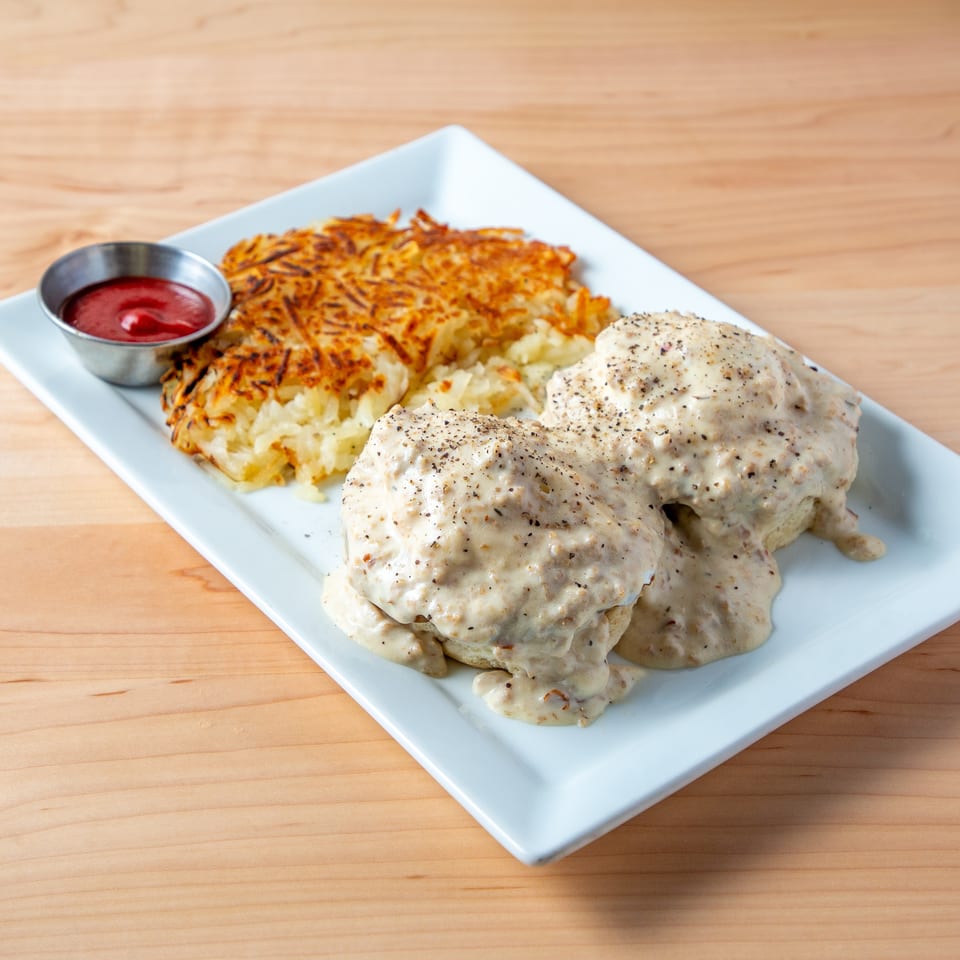 Biscuits and gravy with hash browns and a side of ketchup on a white rectangular plate.