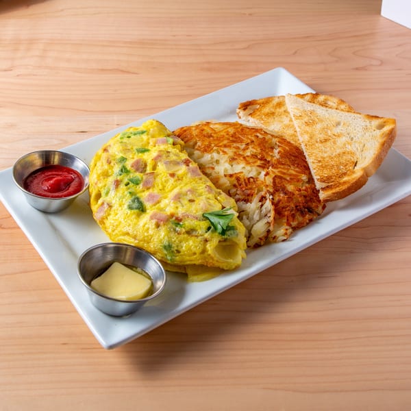 Ham and cheese omelet with hash browns, toast, ketchup, and butter on a white plate.