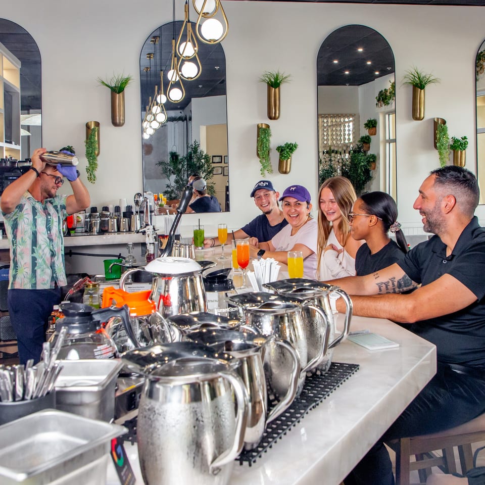 A bartender shakes a drink for four smiling customers seated at a bright, modern bar.