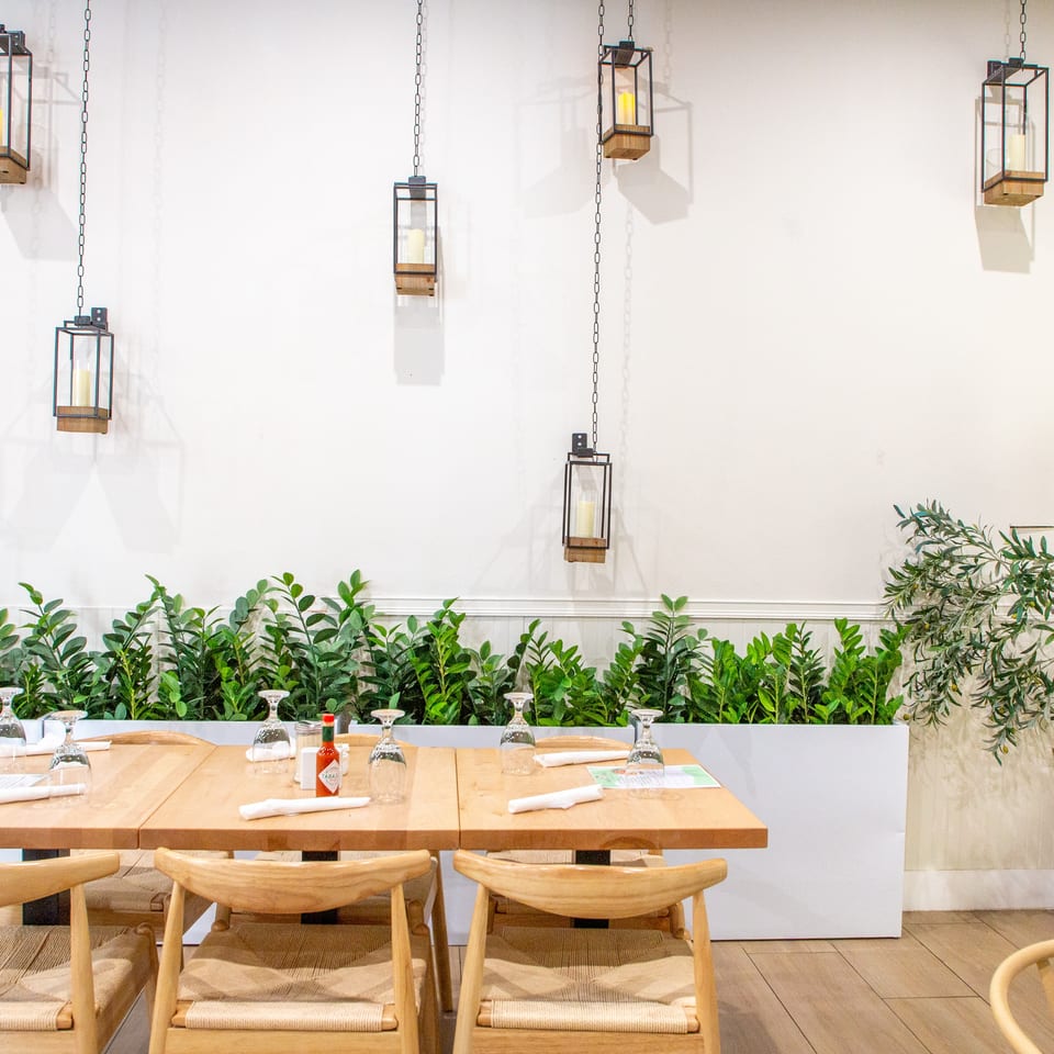 Restaurant interior with light wooden tables, chairs, green plants, and hanging candle lanterns.
