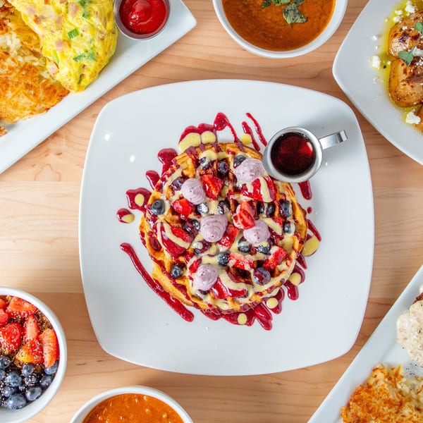Overhead view of a waffle with berries and cream, surrounded by various breakfast dishes on a wooden table.