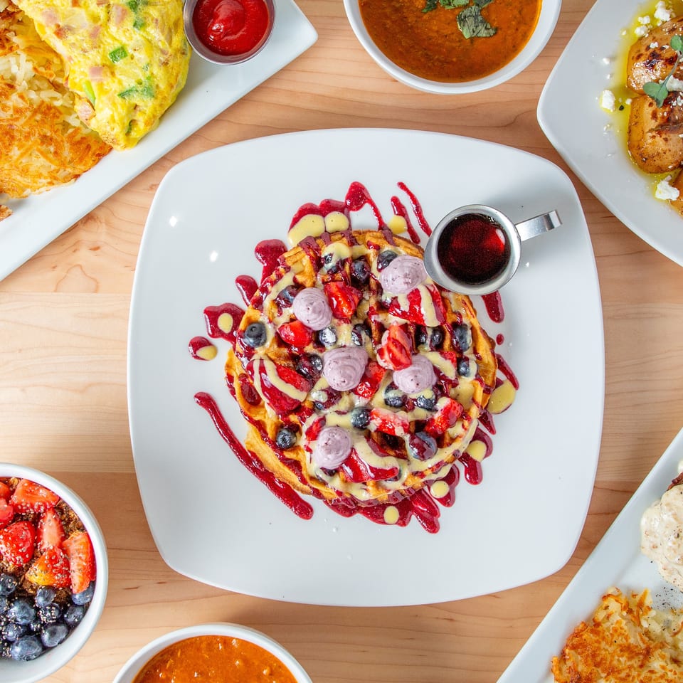 Overhead view of a waffle with berries and cream, surrounded by various breakfast dishes on a wooden table.
