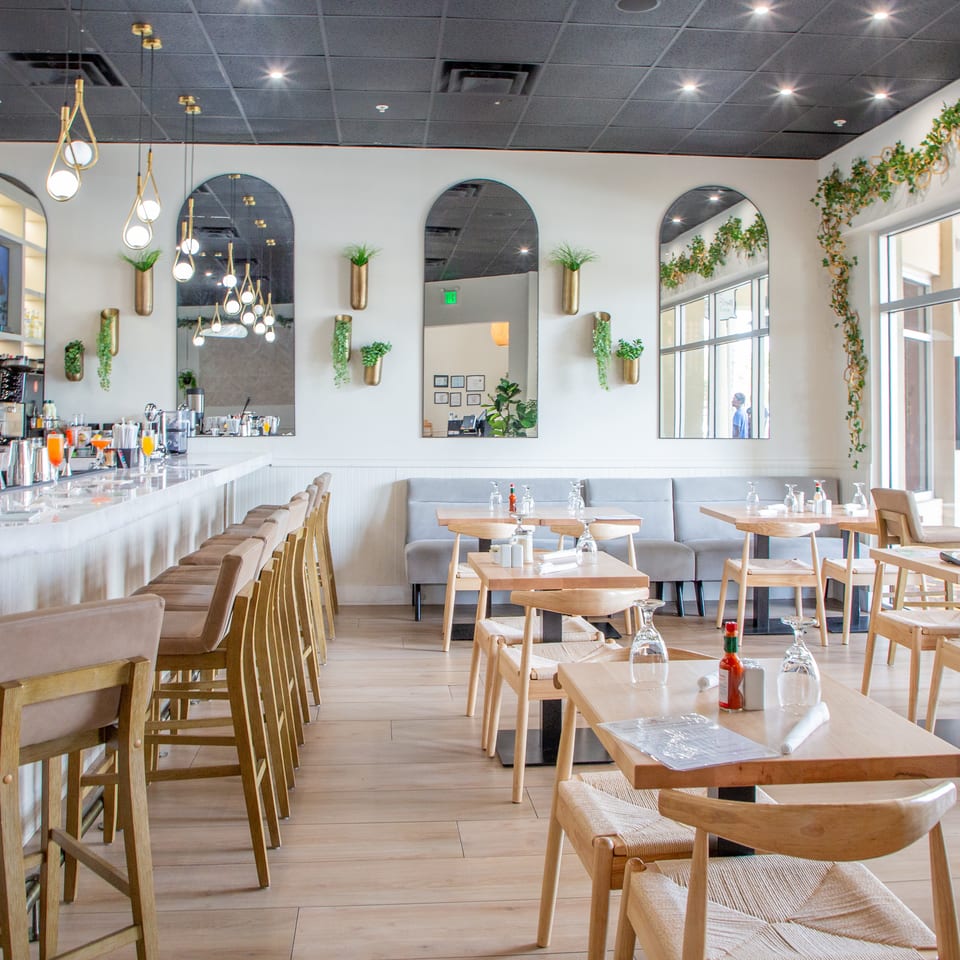 Bright restaurant interior with a long bar, light wood tables, gray booths, and arched mirrors with plants.