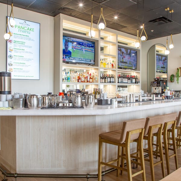 A modern bar area with a white marble counter, bar stools, liquor shelves, TVs, and a pancake menu.