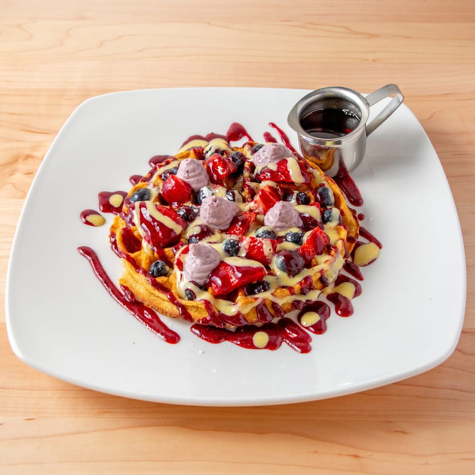 Waffle topped with strawberries, blueberries, purple cream, and syrup on a white plate.