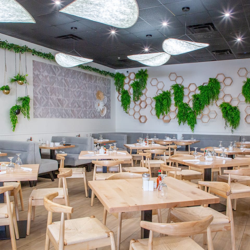 Bright, modern restaurant interior with light wood tables, gray booths, and green plant decor on white walls.